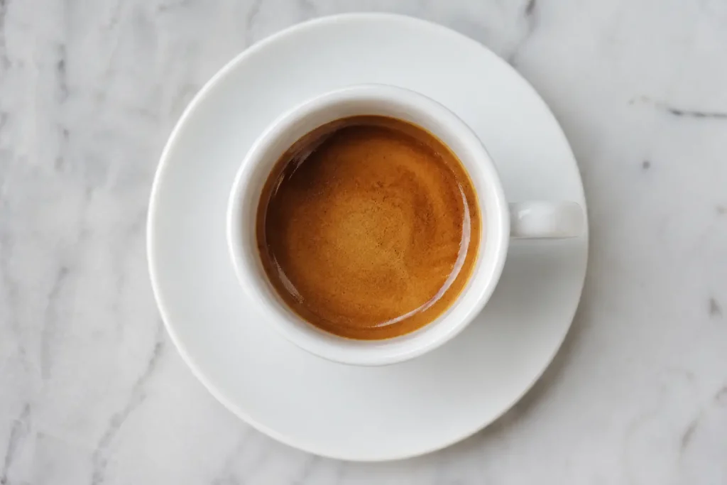 Filiżanka espresso z kawy Bourbon na białym spodku — ciemnobrązowy napar z widoczną złotą cremą, jasne naturalne tło
