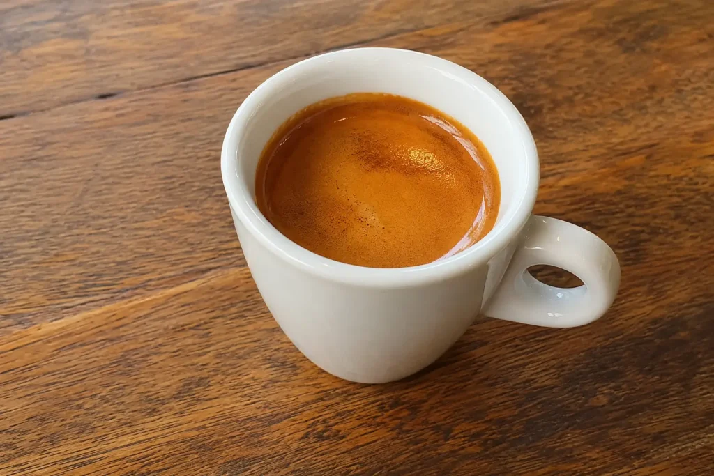 Filiżanka brazylijskiego espresso na drewnianym stole — ciemnobrązowe espresso z wyraźną złotą cremą w białej filiżance, naturalne ciepłe oświetlenie