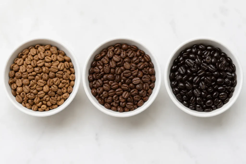 Trzy rzędy ziaren kawy obok siebie — od lewej jasnobrązowe (light roast), przez średniobrązowe (medium roast) po ciemnobrązowe z oleistą powierzchnią (dark roast), widok z góry na białym tle