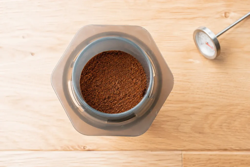 Widok z góry na komorę AeroPress ze świeżo zmieloną kawą i termometrem obok, na jasnym drewnianym blacie.